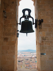 Obraz premium A big old medieval wrought iron bell hanging in the bell tower of the Cathedral of Segovia and city landscape at the background.
