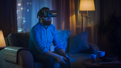 Fototapeta premium Man Sitting on a Couch in His Living Room Wearing Plays Video Games on His Console While Wearing Virtual Reality Headset.