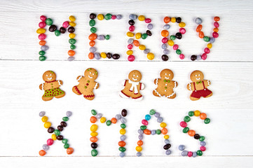 Naklejka premium Inscription of colorful sweet candy on a white wooden table - Merry Christmas / Merry Xmas! Funny painted gingerbread men for the new year. Boys and girls from cookies.
