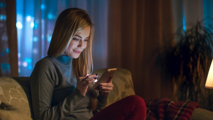 Beautiful Young Woman Sitting on Her Sofa in a Living Room. She Uses Smartphone with Interest. It's Evening, Big City is Seen in the Window.
