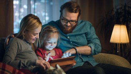 Father, Mother and Little Daughter Reading Children's Book on a Sofa in the Living Room. It's...