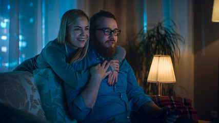 In the Evening Man Sitting on a Sofa Playing Videogames, His Spouse Comes in and Hugs Him.