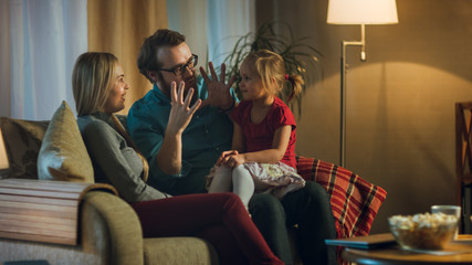 Fototapeta premium In the Evening Family Spends Time Together Sitting on a Couch in a Living Room. Little Girl Sits on Her Father's Laps and Parents Tickle Her.
