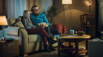 Obraz premium In the Evening Man and Woman are Sitting on the Sofa Watching TV and Eating Popcorn. Living Room is Cozy.