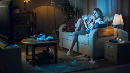 In the Evening Heartbroken Girl Sitting on a Sofa, Crying, Using Tissues, Eating Ice Cream and Watching TV. Her Room is in Mess.