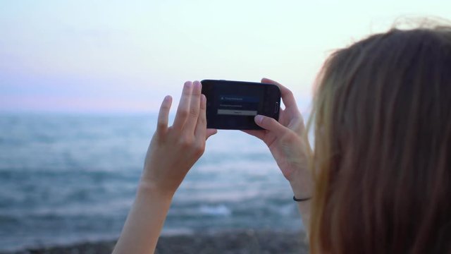 Young hipster woman making photo on smartphone by the sea shore