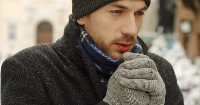 Close up of the sad handsome man in a hat and warm clothes thinking and warming his hands in gloves on the street with Christmas decorations. Portrait. Outdoor