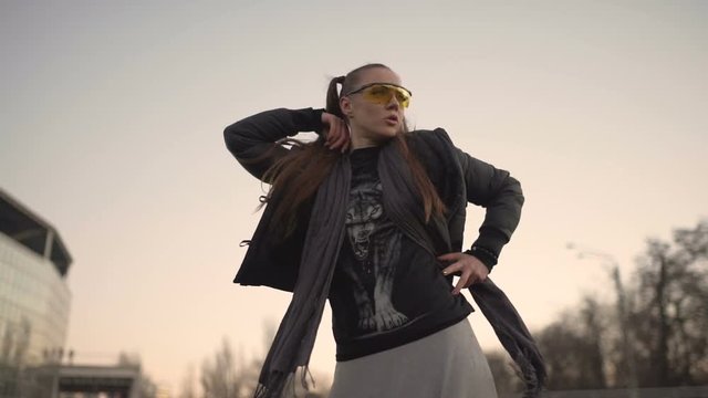 Attractive young hipster woman in a stylish look dancing freestyle on urban background at sunset or dusk
