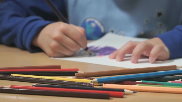 Close-up of hands of unidentified little boy in blue sweater sitting at table drawing pictures using multicolored pencils in kindergarten