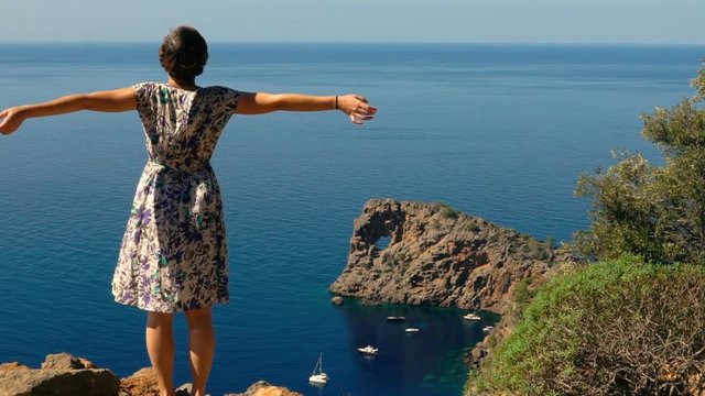 Young happy woman reaches top of the mountain cliff and raises her arms successfully against the beautiful ocean landscape