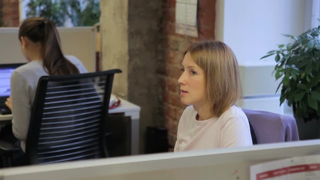 Female specialist talks sitting in workplace in leading company. Woman is speaking actively, looking ahead and gesticulating with hands, then turns body and looks attentively at pc screen. Attractive