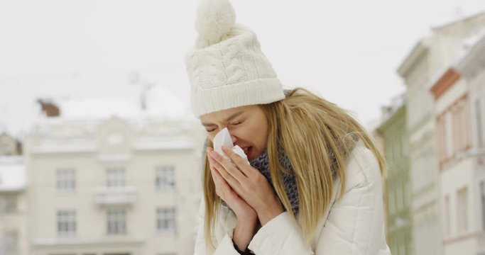 Ill young blonde woman blowing her nose int he napkin on the city square on winter day. Close up. Portrait. Outdoors