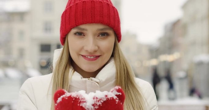 Portrait of the beautiful blonde woman in red hat and gloves showing snow in hands palms to the camera and blowing it on the street of city on winter day. Close up. Outside