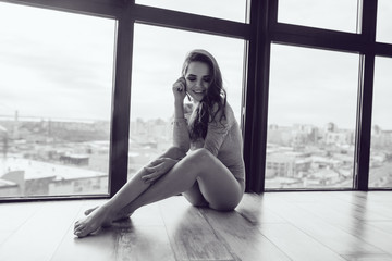 Sexy woman in gray bodysuit sits on the floor near the panoramic window.