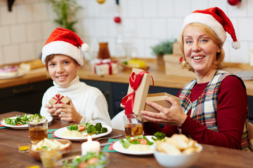 Cheerful grandmother and grandson with gifts sitting by festive table on Christmas evening
