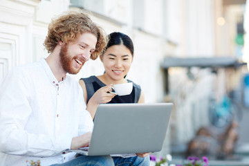 Asian girl and Caucasian guy with laptop watching curious video in the net in urban environment