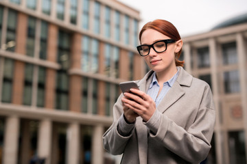 Young contemporary woman reading notification in her smartphone while walking in urban environment