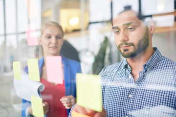 Obraz premium Businessman looking at one of notepapers on transparent office board with colleague near by