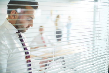 Fototapeta premium Businessman with touchpad standing by transparent office wall with jalousie and networking