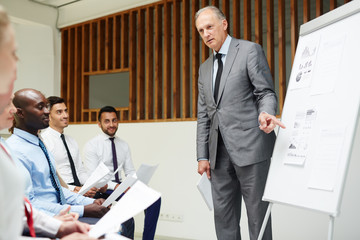 Confident and experienced financier pointing at one of papers on whiteboard and explaining...