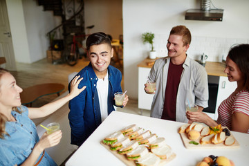 Attractive young man with drink looking at one of girls during talk at party