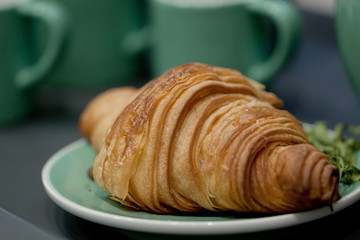 appetizing croissant on a plate and on a background of green cups, will help you to quickly have a snack in a cafe