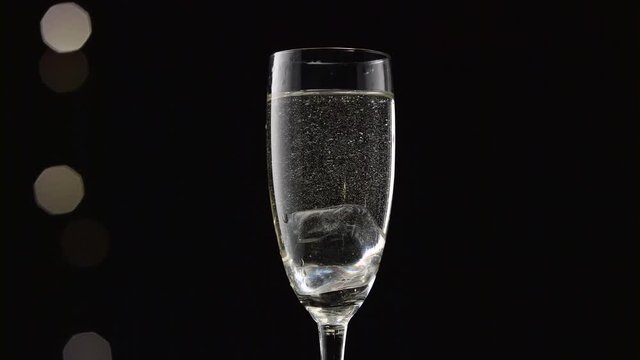 Cube of ice falls into a glass of champagne. Bokeh blinking black background. Close up