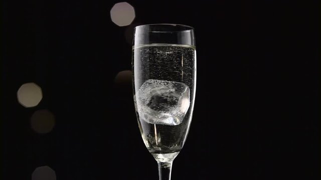 Cube of ice swirls in a glass of champagne. Bokeh blinking black background. Close up