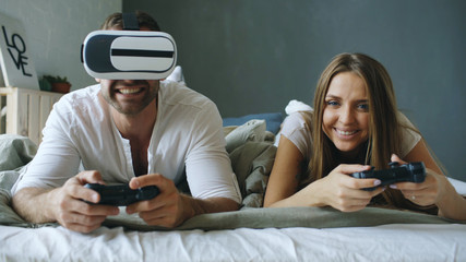 Young couple lying in bed play video games with controller and VR headset