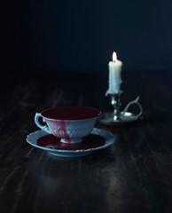 White tea cup in dark blood on dark wooden table with burning candle in candleholder.