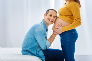 Fototapeta premium I hear him. Positive delighted future father keeping smile on his face and putting hands on the baby belly while looking forward