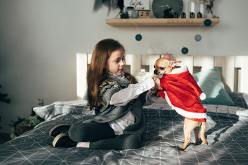A little girl holds in the hands of a cute dog and sits on the bed