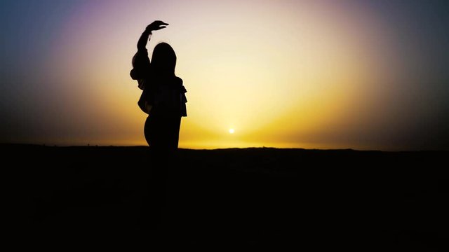 Pretty sexy girl dances in desert with sun on background. Happy beautiful woman is dancing east dance at sunset. Dancer melts, dissolves in sunset