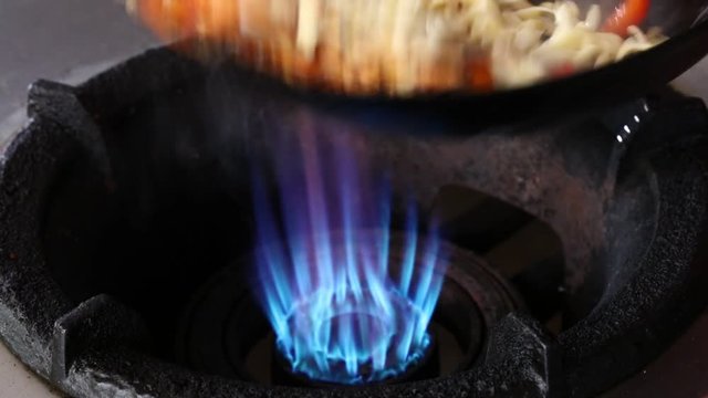 Close up cooking stir fried noodles and vegetables, pepper and carrots, in wok pan on open gas fire, high angle view