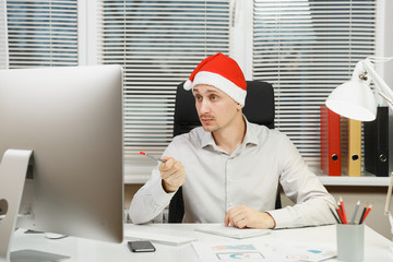 Handsome serious and engrossed business man in shirt, red Christmas hat sitting at the desk,...
