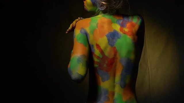 A small woman in a multi-colored body art looks back and covers her breast with her hand