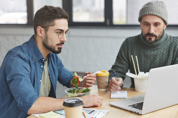 Young handsome male vegeterian eats healthy salad at work place, looks attentively at laptop, sits near his mature colleague, discuss together illustrations for project, browse web site use internet