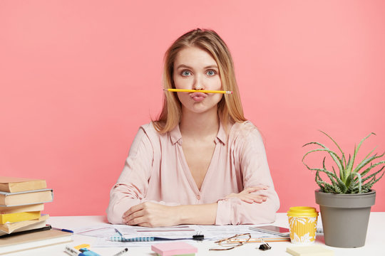 Funny female student foolishes as being exhausted of studying, pouts lips and keeps pencil, feels bored, wants to have entertainment instead of learning material, isolated on pink background
