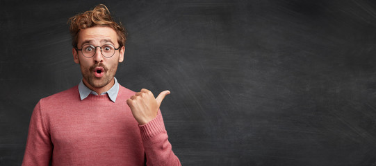 Horizontal shot of shocked male student with stylish hairdo, stares through spectacles, being surprised to indicate something at copy space for your promotional text or hearder. Advertising concept