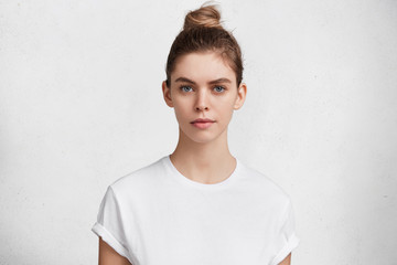 Serious pleasant looking woman with dark blonde hair tied in bun, wears casual white t shirt going...