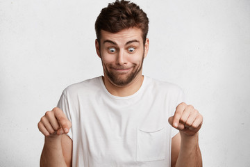 Funny surprised bearded male model looks with bugged eyes down and indicates as shows something, sees comic things, isolated over white concrete wall. Positive amazed young man poses in studio © WHstudio Leushin N