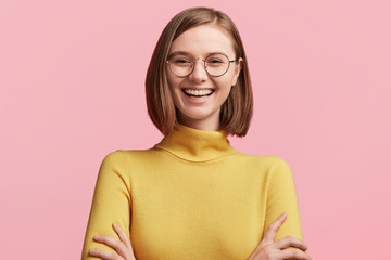 Obraz premium Cheerful female student has fun during break in colleague, laughes at jokes of groupmates, keeps hands crossed, wears round optical glasses and yellow turtleneck sweater. Positive emotions concept