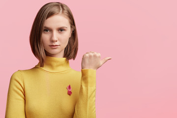 Horizontal shot of serious female has attractive appearance wears red ribbon denotes AIDS virus, support people with immune deficiency syndrome, points with thumb at blank copy space for advertisment