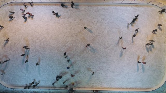 People are skating on illuminated ice rink in the evening. Aerial vertical top-down view. Establishing revealing shot. Drone is flying upward.