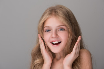 Cannot believe. Close-up portrait of surprised lovely girl is standing and smiling while looking at camera with astonishment. She is holding hands by her face. Isolated background