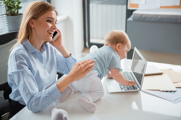 Side view beaming young mom taking care of kid. He creeping on desk. Cheerful woman telling by phone. Profession and child concept