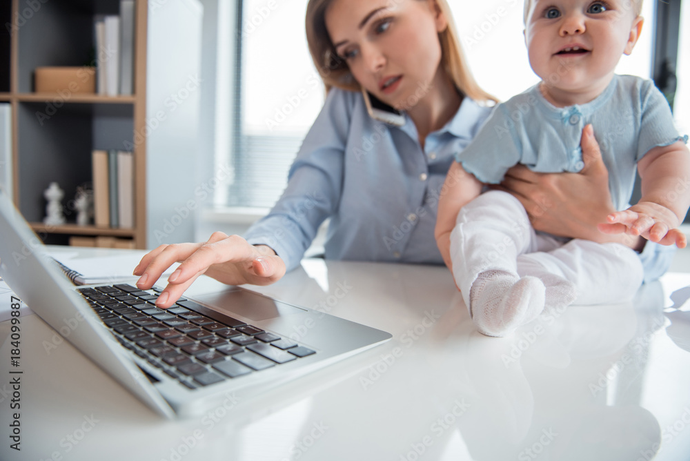Poster serene woman working in digital device and talking by mobile while keeping smiling kid on table in m