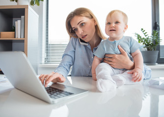 Portrait of serene mother typing in laptop and speaking by phone while holding little child on desk in room. Infant at work concept