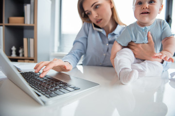 Serene woman working in digital device and talking by mobile while keeping smiling kid on table in modern office. Labor and baby concept