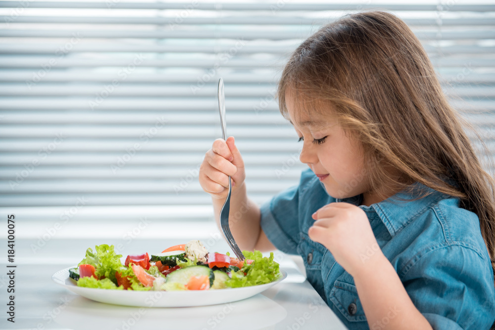 Wall mural profile of cute little asian girl having healthy breakfast at home. she is looking in plate of salad